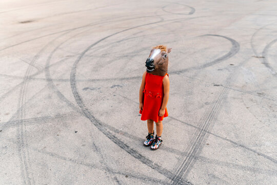 Little girl with a horse's head and a red dress, standing on asphalt with tire tracks
