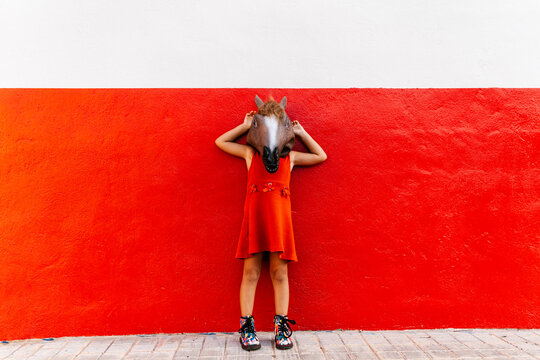 Little girl with a horse's head and a red dress Lening on a red and white wall