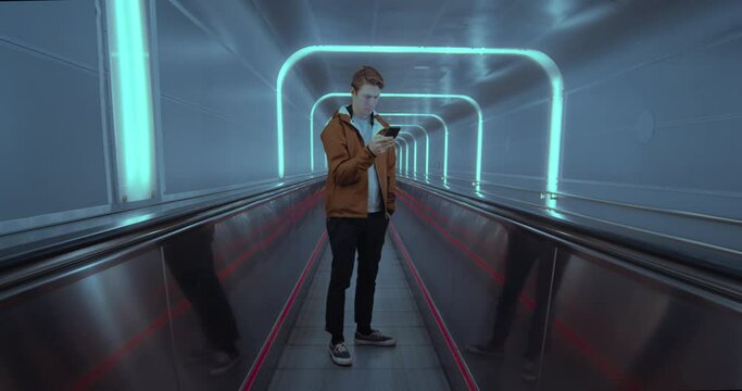 Young modern man distracted by smartphone in futuristic urban environment. Millennial generation z hipster travelling on moving walkway in airport or subway. Modern lifestyle concept