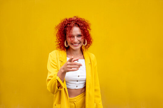 Happy young woman with red curly hair pointing with finger in front of yellow wall