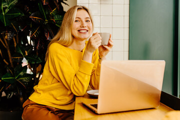 Blond girl with cup of coffee in trendy clothes sitting near the window counter surrounded with green plants, holding laptop on table and gazing with cute smile at camera.