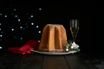 Pandoro, a typical Italian cake for Celebrating Christmas. The cake is on a black wooden table with a glass of sparkling wine, Christmas decorations and a bokeh effect on a dark background.