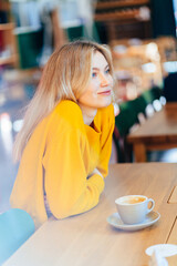 Portrait of blond woman in yellow pullover sits with cup of coffee or tea at the beautiful loft...