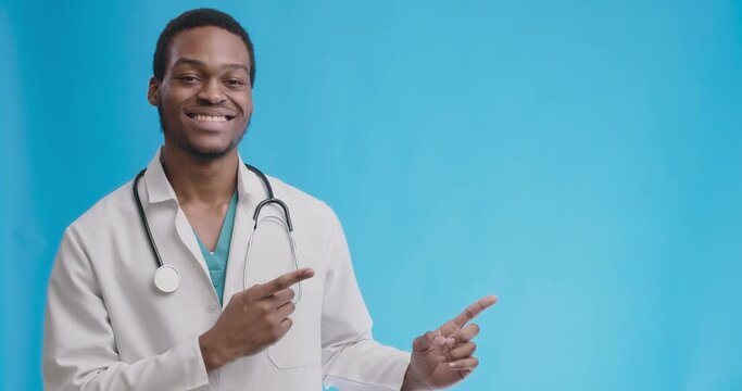 Cheerful african american professional doctor showing empty space for mock up, pointing aside aon blue studio background