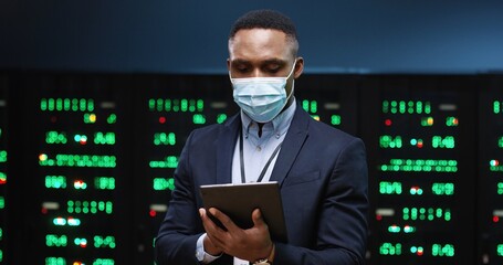 Close up portrait of handsome African American man wearing medical mask standing among servers with tablet and typing on screen and checking big data processors. Male analytic working in data storage