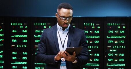 Close up portrait of African American young male IT technician specialist in glasses tapping on tablet device while standing in big data center running diagnostics of processors in data storage
