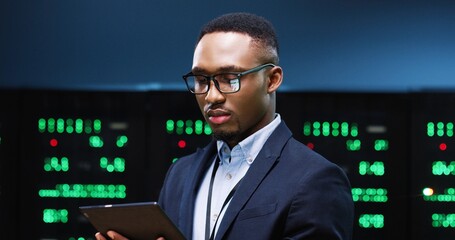 Close up of handsome African American young businessman IT technician specialist in glasses browsing on tablet device while standing in server room running diagnostics of processors. Big data center