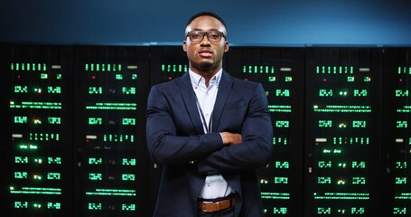 Portrait of handsome African American serious male IT security engineer standing in big data center with server racks and looking at camera. Man analytic in glasses at work in server room Tech concept