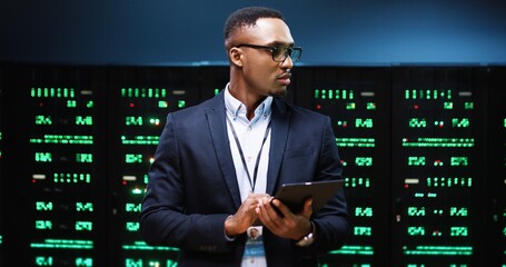 Close up portrait of African American young male IT technician specialist in glasses tapping on tablet device while standing in big data center running diagnostics of processors in data storage