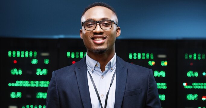 Close Up Portrait Of Cheerful African American Handsome Male System Administrator In Good Mood Stands In Big Data Center With Server Racks And Smiling. Man IT Professional In Glasses In Server Room