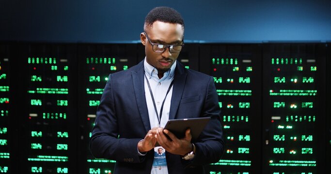 Close up portrait of African American young male IT technician specialist in glasses tapping on tablet device while standing in big data center running diagnostics of processors in data storage