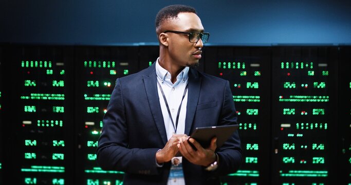 Close up portrait of African American young male IT technician specialist in glasses tapping on tablet device while standing in big data center running diagnostics of processors in data storage