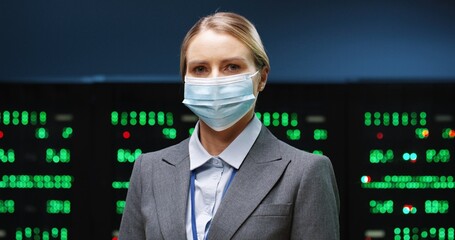 Close up portrait of beautiful Caucasian woman professional security engineer in medical mask standing in datacenter and looking at camera. Young happy female administrator in data storage Server room