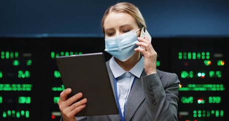 Close up portrait of pretty female system administrator in mask standing in data storage room working on digital security of processors and typing on tablet while talking on cellphone. Big data