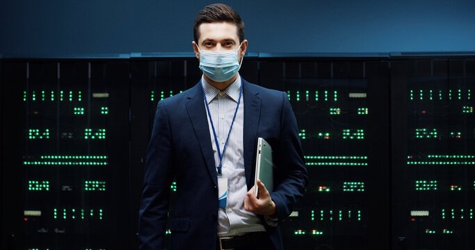 Portrait of Caucasian male security engineer in mask standing in datacenter checking security of processors network with digital information. Handsome man work in big data storage Technologies concept