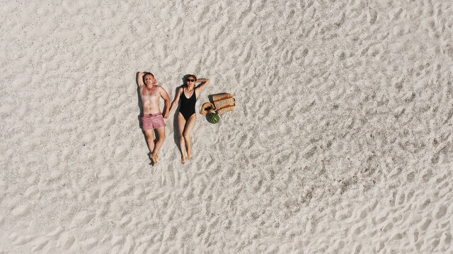 Aerial View Of A Young Couple Lying On The White Beach Sand. Man And Woman In Swimwear Spend Time Together And Travel Through The Desert