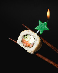 Sushi on black background. Philadelphia maki roll piece between chopsticks with green star shaped burning candle. Christmas and New Year celebration party. Creative japanese food. Faded, close up