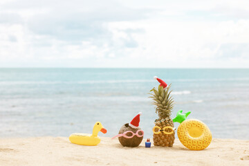   funny attractive pineapples and coconut in stylish sunglasses on the sand against turquoise sea. Wearing christmas hats. 