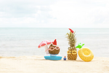 funny attractive pineapples and coconut in stylish sunglasses on the sand against turquoise sea. Wearing christmas hats. 