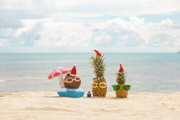  funny attractive pineapples and coconut in stylish sunglasses on the sand against turquoise sea. Wearing christmas hats. 