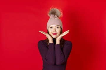 close up of a smiling young woman in a knitted winter hat and hands near her face, isolated on a red background.