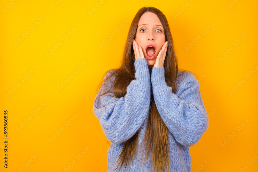 Wall mural Scared terrified Young beautiful Caucasian woman wearing blue sweater against yellow wall shocked with prices at shop, People and human emotions concept - Wall murals