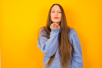 Young beautiful Caucasian woman wearing blue sweater against yellow wall looking at the camera blowing a kiss with hand on air being lovely and sexy. Love expression.