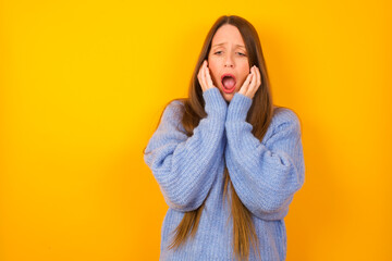 Stupefied Young beautiful Caucasian woman wearing blue sweater against yellow wall expresses excitement and thrill, keeps jaw dropped, hands on cheeks, has eyes popped out