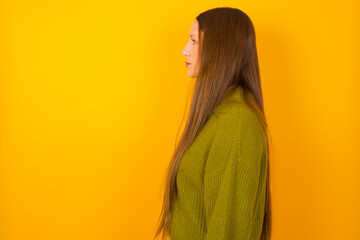 Young beautiful Caucasian woman wearing green sweater against yellow wall looking to side, relax profile pose with natural face with confident smile.