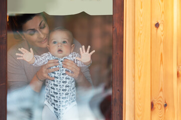 Happy mother and her baby boy looking throw the window and playing in warm home on winter day. Christmas time.