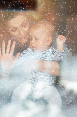 Happy mother and her baby boy looking throw the window and playing in warm home on winter day. Christmas time.