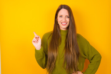 Young beautiful Caucasian woman wearing green sweater against yellow wall looking at camera indicating finger empty space sales