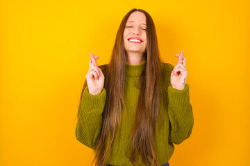 Young beautiful Caucasian woman wearing green sweater against yellow wall has big hope, crosses...
