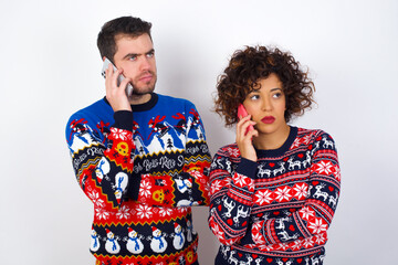 Sad Young couple wearing Christmas sweater standing against white wall talking on smartphone. Communication concept.