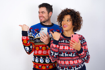 Young couple wearing Christmas sweater standing against white wall using and texting with smartphone  pointing and showing with thumb up to the side with happy face smiling