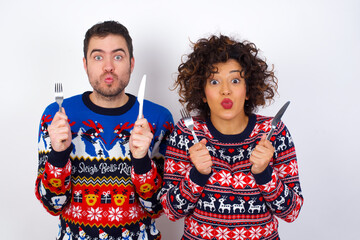 Model hungry Young couple wearing Christmas sweater standing against white wall holding in hand fork knife want tasty yummy pizza pie