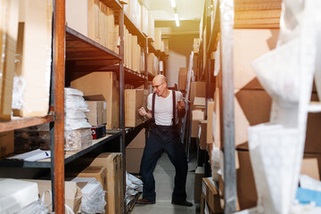 Fototapeta na wymiar Happy funny warehouse male worker dancing between tall rows of shelves. He has bold head, wearing suspenders and glasses.