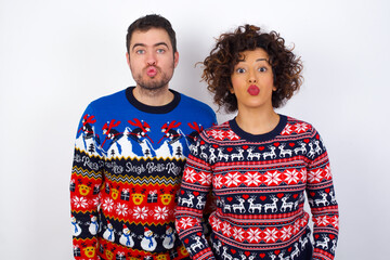 Shot of pleasant looking Young couple wearing Christmas sweater standing against white wall, pouts lips, looks at camera, Human facial expressions