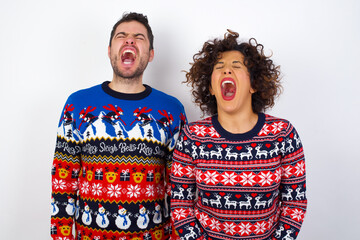 Young couple wearing Christmas sweater standing against white wall angry and mad screaming frustrated and furious, shouting with anger. Rage and aggressive concept.