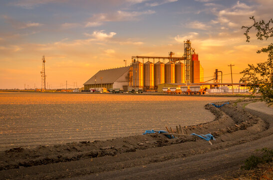 A large grain elevator is standing at a distance with a plowed field and small water stream in front. Sunset with orange skies and clouds. 