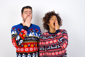 Young couple wearing Christmas sweater standing against white wall being tired and yawning after spending all day at work.