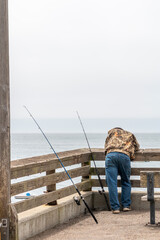 A man is on a pier leaning over a wooden railing looking down at the ocean. Two fishing poles are to his left and have lines down into the water. A small white boat can be seen between the wooden rail