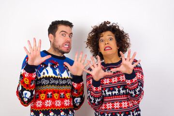 Young couple wearing Christmas sweater standing against white wall afraid and terrified with fear expression stop gesture with hands, shouting in shock. Panic concept.