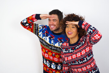 Young couple wearing Christmas sweater standing against white wall very happy and smiling looking far away with hand over head. Searching concept.