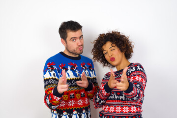 Joyful Young couple wearing Christmas sweater standing against white wall wink and points index fingers at camera, chooses someone, makes finger gun pistol