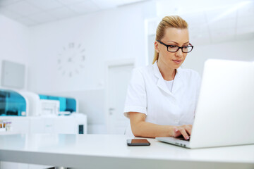 Middle aged attractive lab assistant with eyeglasses sitting in laboratory and typing on laptop...