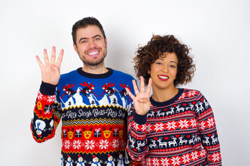Young couple wearing Christmas sweater standing against white wall showing and pointing up with fingers number four while smiling confident and happy.