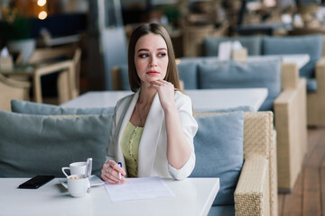 Young woman in white jacket working in cafe with papers, drinking coffee