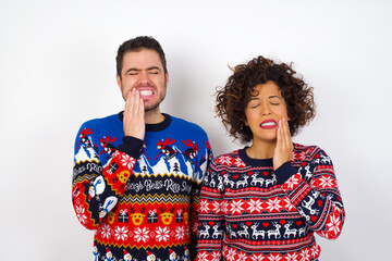 Young couple wearing Christmas sweater standing against white wall touching mouth with hand with painful expression because of toothache or dental illness on teeth.