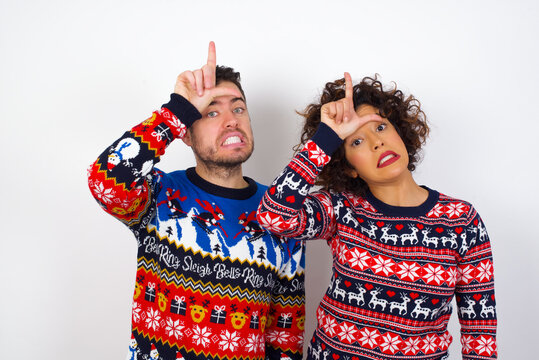 Young couple wearing Christmas sweater standing against white wall making fun of people with fingers on forehead doing loser gesture mocking and insulting.